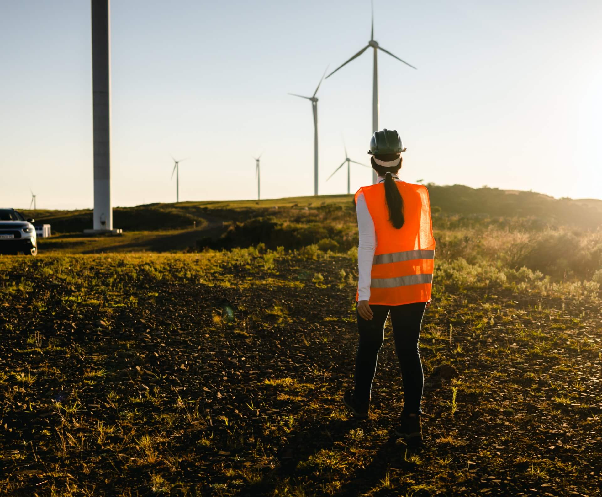 A woman wearing an orange vest poses in front of several wind turbines